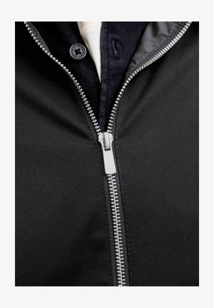 Zbliżenie srebrnego zamka błyskawicznego na czarnej kurtce z ciemną koszulą i białą podkoszulką widoczną pod spodem.