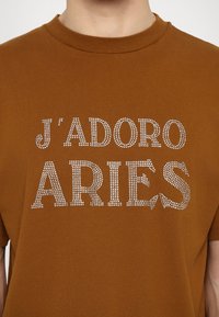 Brown cotton t-shirt featuring "J'ADORE ARIES" in silver rhinestones. Crew neck design, short sleeves, and a smooth texture.