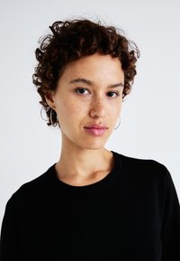 Svart tröja med ribbad rund halsringning, bärs av en person med kort, lockigt hår. Framträdande fräknar och små hoop-örhängen.
