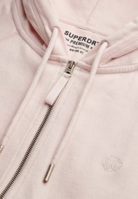 Sweat à capuche Superdry rose clair avec fermeture éclair en métal et cordons de serrage brandés, avec logo brodé sur la poitrine.