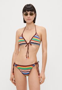 Ensemble de bikini au crochet présentant un motif rayé multicolore avec des accents rouges. Haut triangle et bas à nouer sur les côtés, ornés de petits anneaux décoratifs.
