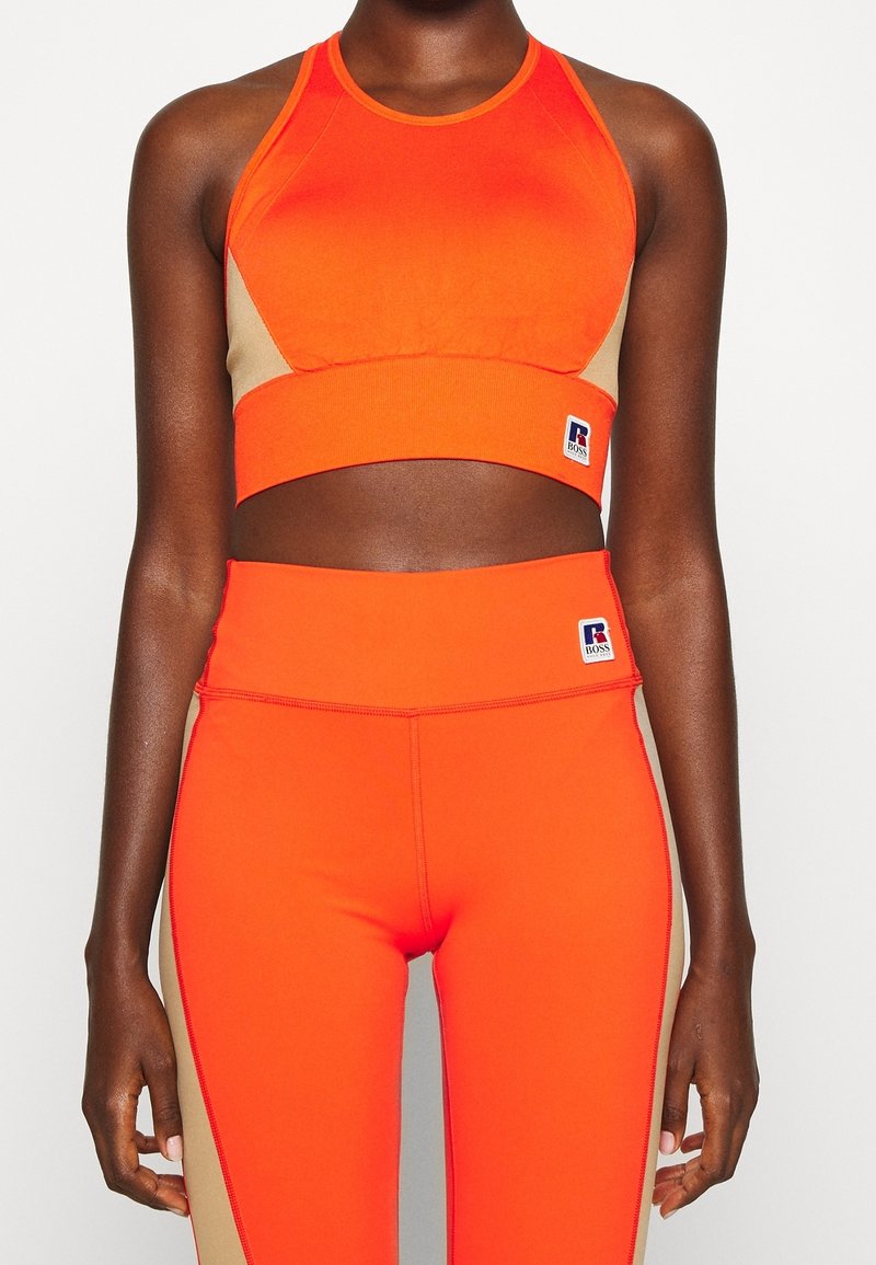 Brassière de sport orange vif et leggings taille haute, avec des accents couleur beige, un tissu extensible et une étiquette logo sur la ceinture.