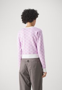 Suéter recortado en rosa claro con un patrón de tablero de ajedrez blanco, con mangas largas y un dobladillo blanco acanalado. Pantalones grises con bolsillos.