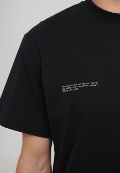 Tricou din bumbac organic negru, cu mâneci scurte. Are un text imprimat alb pe față, care conține instrucțiuni de îngrijire și detalii despre material.