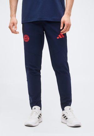 Man wearing navy blue sports pants with red Adidas and FC Bayern Munich logos, paired with white Adidas sneakers, standing on a white background.