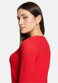 Czerwony sweter o fakturowanej powierzchni z długimi rękawami, z okrągłym dekoltem i dopasowanym krojem, widoczny z boku.