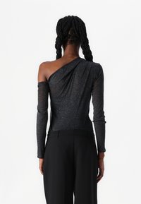 Schwarzes Ein-Schulter-Top aus schimmerndem, strukturiertem Stoff. Mit langen Ärmeln und einem drapierten Design. Kombiniert mit maßgeschneiderten schwarzen Hosen.