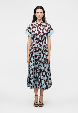 Kvinde stående iført en brun midi-kjole med store lyseblå blomsterprint, korte ærmer, knappet foran og sorte ankelremshæle.