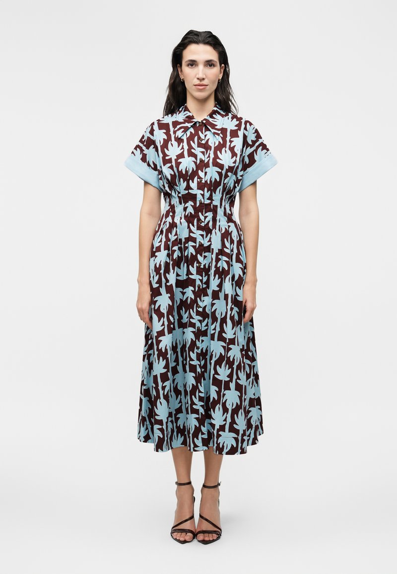 Γυναίκα στέκεται φορώντας ένα καφέ μίντι φόρεμα με μεγάλα φωτεινά μπλε λουλουδένια σχέδια, κοντά μανίκια, κουμπωτό μπροστά και μαύρα τακούνια με λουράκι στον αστράγαλο.