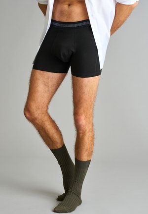Sorte bomulds-boxershorts med en tætsiddende design, forsynet med en grå mærkevare-elastik, båret med olivengrønne ribbede strømper.