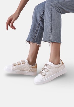 Jambes en jean bleu raccourci portant des baskets blanches avec imprimé léopard doré et trois bandes velcro dorées, posées devant un fond uni.