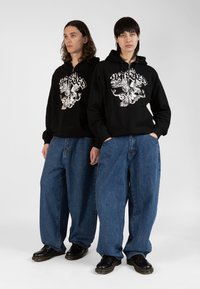 Black zip-up hoodies with white graphic designs, paired with loose-fitting blue jeans and black shoes. Flat background, focused on apparel.