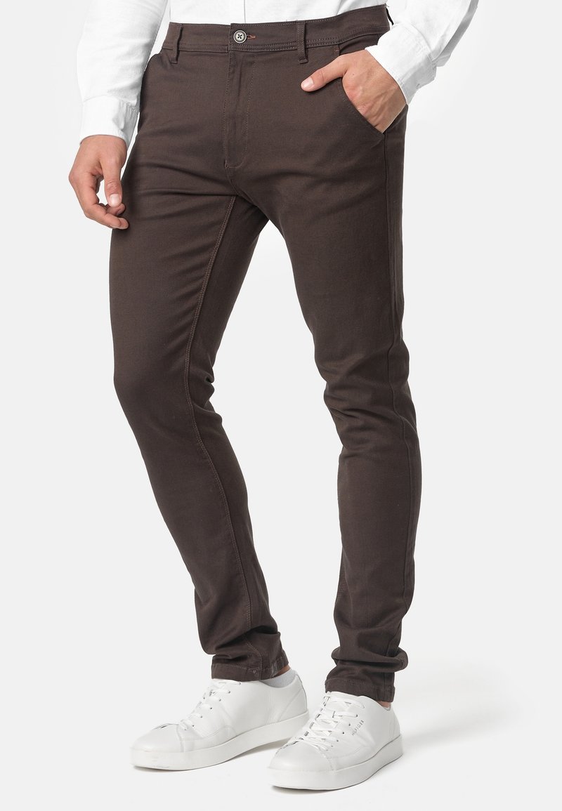 Homme portant un pantalon slim marron foncé, une chemise blanche à manches longues et des baskets blanches, une main dans la poche.
