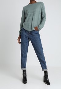 Hellgrüner, locker sitzender Strickpullover, kombiniert mit hochgeschnittenen blauen Jeans und schwarzen Stiefeletten.