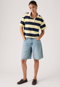 Νεαρή γυναίκα που φοράει μπλούζα πόλο με κίτρινες και ναυτικές ρίγες, ανοιχτό μπλε τζιν σορτς μέχρι το γόνατο και καφέ παπούτσια τύπου boat shoes, με τα χέρια στις τσέπες.