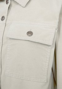 Ljust beige kord-överdel med knappstängning och en enda ficka på bröstet med tryckknapp. Texturerade ribbor är synliga.