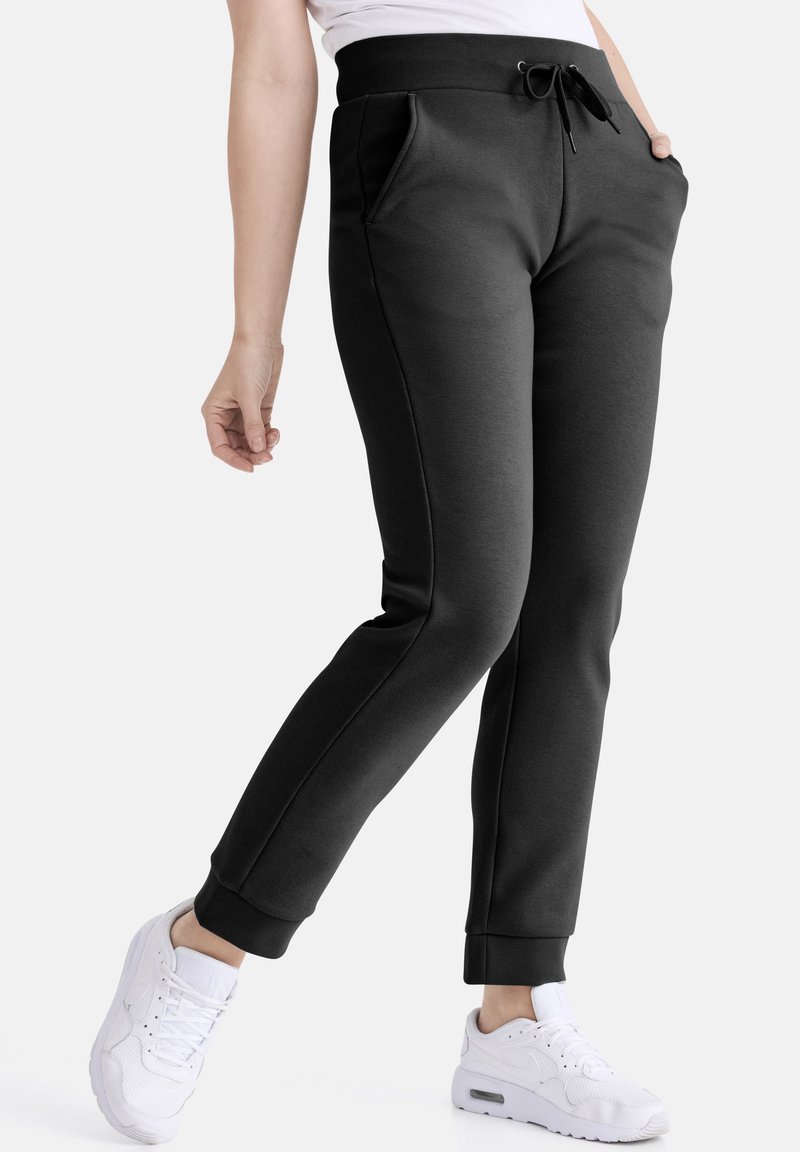 Schwarze Jogginghosen mit elastischem Bund, Kordelzug, Seitentaschen und zugeknöpften Bündchen. Getragen mit weißen Sportschuhen.