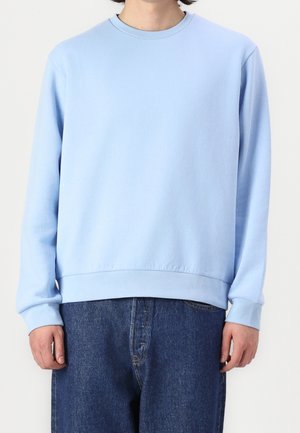 Hombre vestido con un sudadera de cuello redondo de manga larga azul claro y jeans de mezclilla azul oscuro, de pie frente a un fondo blanco liso.