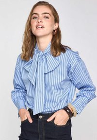 Blusa a righe azzurro chiaro con colletto a fiocco, maniche lunghe e dettagli ai polsini, realizzata in tessuto morbido. Indossata con jeans scuri e braccialetti.