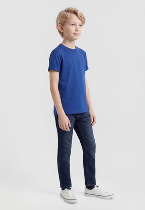 Fiú áll kék pólóban, sötét farmernadrágban és fehér tornacipőben, egyszínű világos szürke háttér előtt.