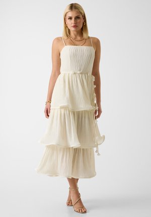 Femme blonde en robe midi crème sans manches à volants, avec fines bretelles, bijoux dorés et talons à lanières, sur fond blanc.