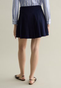 Falda plisada navy con una textura suave, combinada con zapatos de tacón abierto negros con un acento dorado. El fondo es neutro.