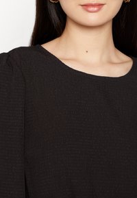 Top preto texturizado com decote arredondado e mangas curtas bufantes, apresentando um padrão sutil e enrugado por todo o tecido.