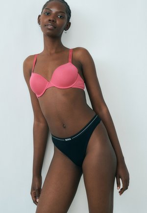 Femeie îmbrăcată cu un sutien roz și chiloți negri cu talie înaltă, având logo-ul „BOSS”, stând în fața unui fundal simplu, gri deschis.