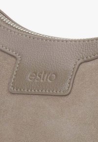 Beżowa torebka zamszowa z jasnobrązową skórzaną metką z wytłoczonym logo "estilo" oraz srebrnym zamkiem błyskawicznym wzdłuż górnej krawędzi.