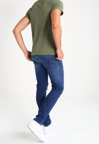 Man standing with back turned wearing olive green t-shirt, blue jeans, and white sneakers against a white background.