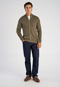 Olijfgroene vest met rits en een getextureerd ruitpatroon, gecombineerd met donkerblauwe spijkerbroek en bruine schoenen met roze veters.