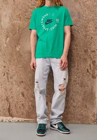 Vihreä puuvillapaita, jossa on ympyrän muotoinen Nike-logo, yhdistettynä vaalennettuihin kulutettuihin farkkuihin ja vihreä-mustiin lenkkareihin, joissa on valkoisia yksityiskohtia.