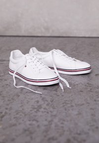 Sneakers bianche in tela con accenti rossi e navy sulla suola, punta tonda e design classico con lacci. Lacci piatti sistemati accanto alle scarpe.