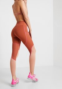 Legginsy z wysokim stanem w kolorze przepalonej pomarańczy z różowymi akcentami, o dopasowanym kroju i siateczkowymi panelami; w zestawie z różowymi butami sportowymi.
