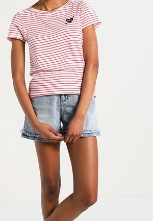 Osoba ubrana w różowo-białą, w paski koszulkę z naszywką serca z napisem „love” oraz jasnoniebieskie krótkie spodenki dżinsowe, stojąca z rękami złożonymi na przodzie.