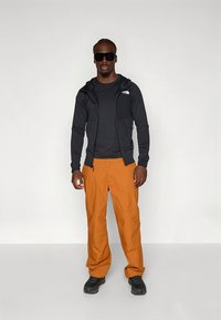 Camisola preta com capuz e fecho éclair, camisa preta, calças laranja e sapatos pretos. Homem em pé sobre um fundo neutro, usando óculos de sol.