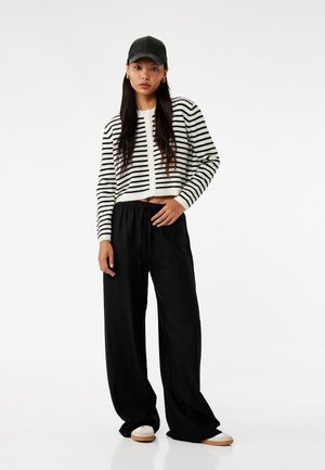 Gestreepte zwart-witte cropped sweater, losse zwarte broek en een zwarte pet, met een zachte textuur en een relaxte pasvorm.