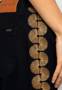 Pantalones de mezclilla negros con un patrón decorativo bordado en oro a lo largo del costado, con una etiqueta de cuero que muestra "THE-ARE" cerca de la pretina.