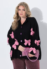 Černý oversized kardigan se růžovými látkovými mašlemi a fialovými knoflíky. V doprovodu malého růžového třpytivého kabelky se středním popruhem.