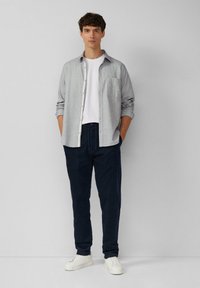 Graues Hemd mit Knopfleiste über einem weißen T-Shirt, kombiniert mit marineblauen Hosen und weißen Sneakers. Das Hemd hat eine Brusttasche und hochgekrempelte Ärmel.