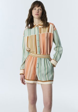 Vrouw staand, gekleed in een button-up, jumpsuit met lange mouwen en verticale strepen in oranje, lichtblauw, beige en bruine tinten.
