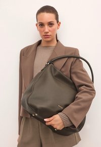 Olívazöld bőr kézitáska lekerekített formával, masszív fekete fogantyúval, diszkrét logórészlettel és fém díszítésekkel az alján.