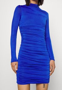 Modré upnuté šaty s dlouhými rukávy a texturovaným řasením po celém obvodu. Mají vysoký límec a hladkou, elastickou látku.