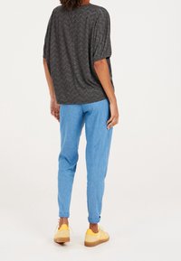 Osoba oblečená v tmavě šedém vršku s chevronovým vzorem, světlých modrých džínech s ohrnutými nohavicemi a žlutých teniskách, stojící zády ke kameře.