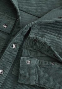 Koszula z materiału w ciemnozielonym korduroju z pionowymi prążkami. Posiada metalowe guziki oraz dwie kieszenie na piersi z klapkami.