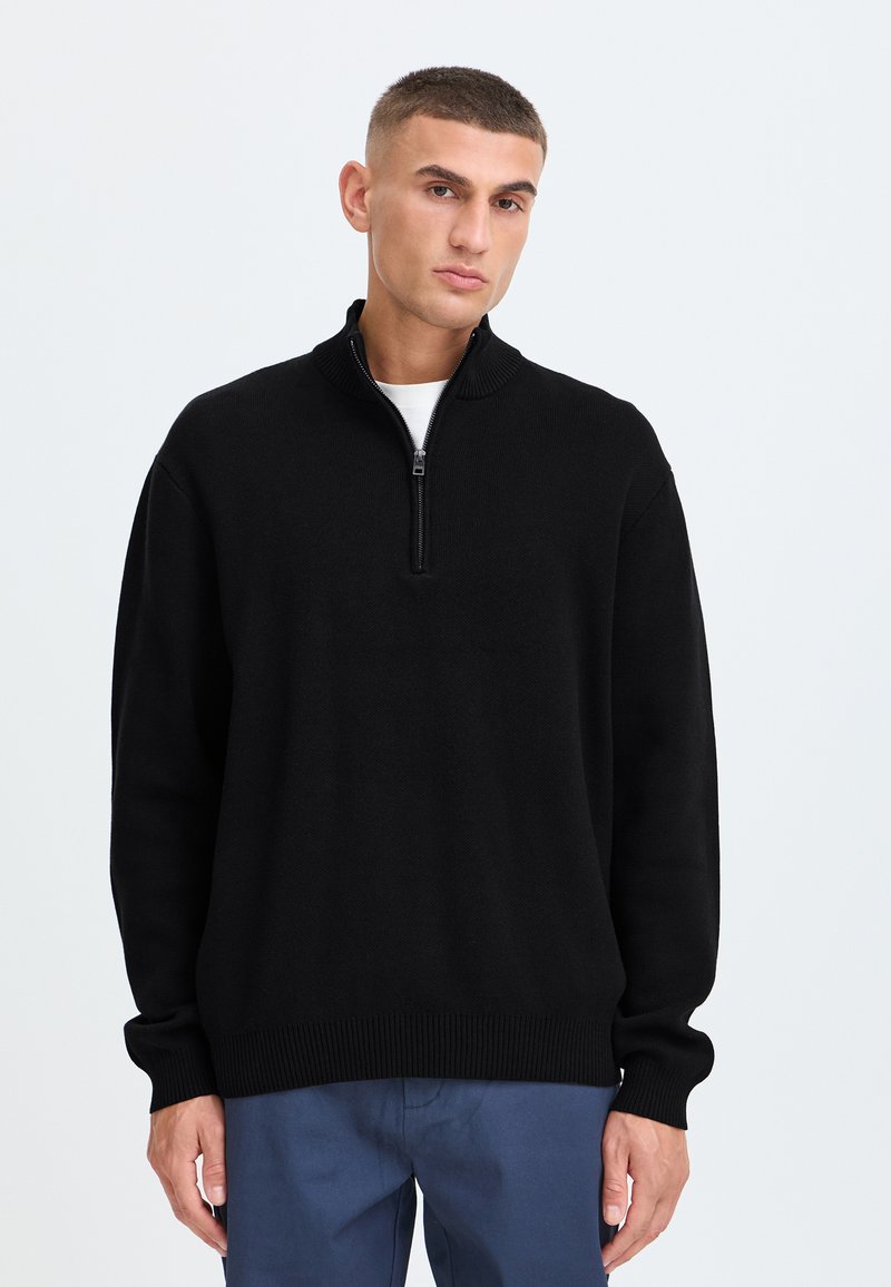 Jeune homme portant un pull zippé noir par-dessus une chemise blanche et un pantalon bleu, debout devant un fond clair et uni.