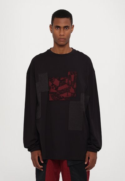 Chemise noire surdimensionnée avec des patchs texturés gris foncé et rouge, présentant un motif détaillé, un col rond et des manches longues.