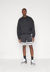 Sort sweatshirt med rund hals og guld logo. Grå sports shorts med hvide striber. Grå sneakers med sorte accenter og hvide snørebånd.