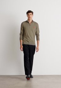 Pullover khaki con zip, realizzato in materiale lavorato a maglia, abbinato a pantaloni neri. Modello con scarpe nere con dettagli rossi, in piedi contro uno sfondo neutro.