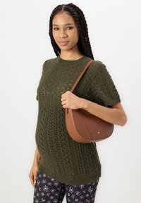 Sac à bandoulière en cuir marron avec une forme incurvée et peu de quincaillerie. Le modèle porte un pull en maille vert et un pantalon noir à motifs.